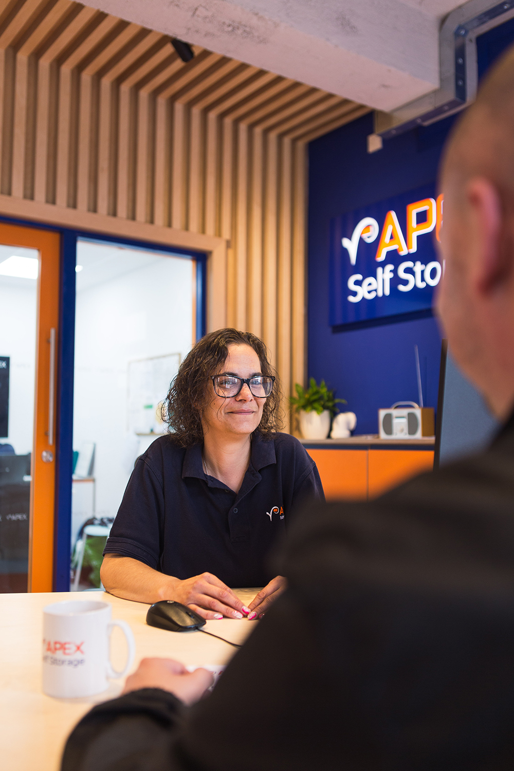 A woman in an Apex Self Storage t-shirt, sitting at a desk and smiling at a customer