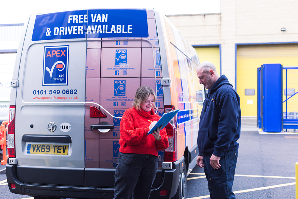 A woman stood next to a removal van with the driver. She is signing a form on a clipboard.